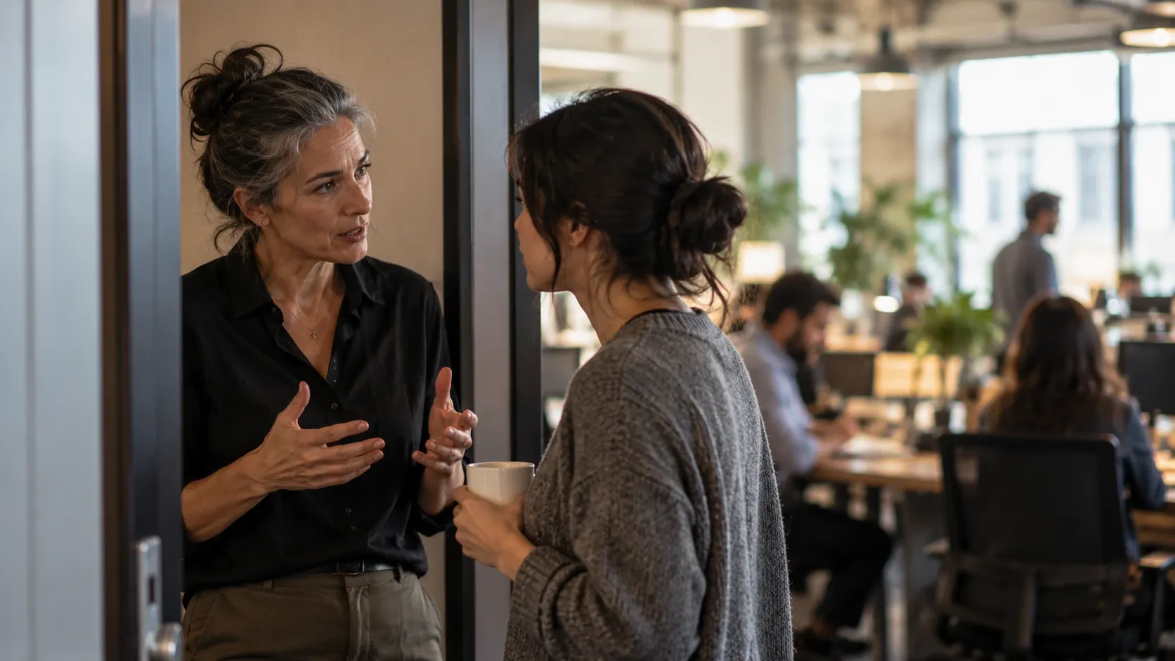 A senior colleague explaining something to a younger coworker in an office doorway while other employees work in the background